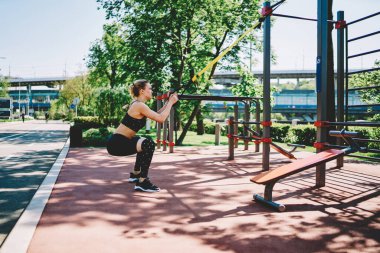 Siyah spor kıyafetli, sarı saçlı atletik bir kadın spor alanında sarı elastik bantla çömelme hareketleri yaparken yazın sokakta spor aletleri ve arka planda büyük yeşil ağaçlarla birlikte.