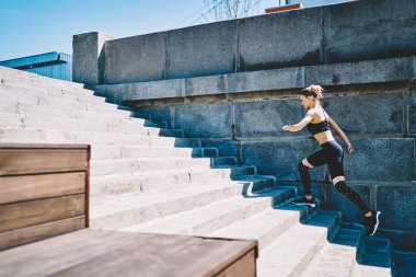 Sıcak güneşli bir günde, sallanan merdivenlerde koşan siyah şık eşofmanlı güçlü sarışın bayan sporcunun yan görüntüsü.