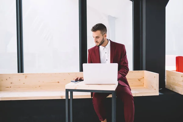 身着深色西装 身穿白衬衫的男性 穿着紧致时髦的紧身衣 坐在木制长椅上 坐在创意的工作空间里 用手机和笔记本电脑查看数据 — 图库照片