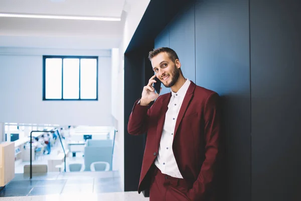 在宽敞的现代办公室里 身穿红色西服 手挽着手 面带微笑的男性 靠着墙看着相机 接电话 — 图库照片