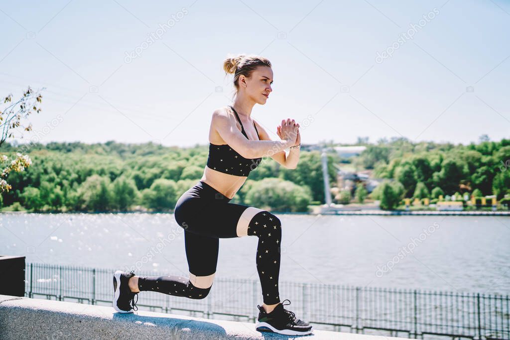 Energetic active woman training and squatting bending leg and folding hands before chest on concrete fence along river in bright day