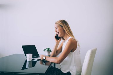 Pozitif kadın resmi giysiler içinde, siyah parlak bir masada otururken, koltuğunda oturup gülerken, hafif modern ofisteki beyaz duvara karşı telefonla konuşurken, yan bakışı.