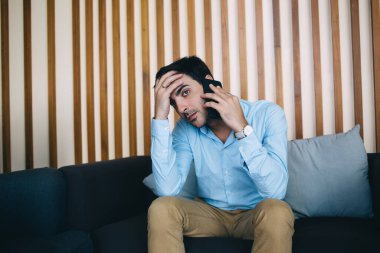 Alnına yaslanmış genç etnik adam modern odada akıllı telefonla konuşurken hayal kırıklığı içinde yüzünü çeviriyor.