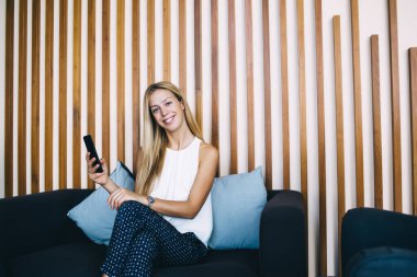 Modern lüks içinde modern lüks içinde oturan neşeli beyaz kadın girişimci cep telefonuyla kameraya bakıyor, modern akıllı telefon kullanan mutlu müreffeh iş kadını.