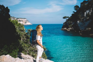 Kafkasyalı kadın Akdeniz kıyılarıyla güneşli doğa manzarasını keşfediyor, Menorca 'ya seyahat eden kadın sırt çantasıyla, nefes kesici manzarası için macera yolculuğuna çıkıyor.