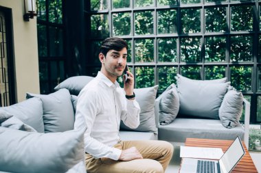 Kafede dizüstü bilgisayarla oturup kameraya bakarken resmi giyinmiş, akıllı telefondan konuşan iyimser erkek liderin yan görüntüsü.