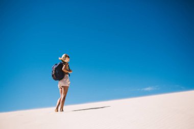 Sırt çantalı çıplak ayaklı sarışın kadının kumlu kumulda dikilip, açık mavi gökyüzüne baktığı arka plan. 