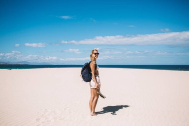 İspanyol plajında güneşlenmek için boş zamanlarında kameraya gülümseyen Kafkasyalı mutlu sırt çantalı kaygısız kadın turist Menorca adasında yaz tatillerinde dinleniyor.