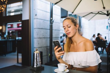 Sosyal medyada cep telefonuyla sohbet eden genç bir kadın bir fincan kahveyle masada oturuyor ve sokak kafesinde dinleniyor.