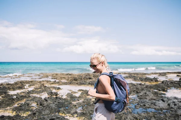 Side view of happy young female traveler in sunglasses with backpack enjoying summer holidays while standing on beautiful rocky seashore on Menorca island