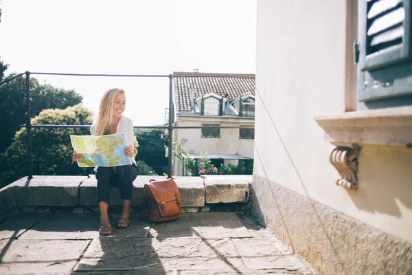 明るい日差しの中で石のテラスのフェンスに座って手の中にリュックと紙の地図を持つ幸せな女性の完全な体離れて見て — ストック写真