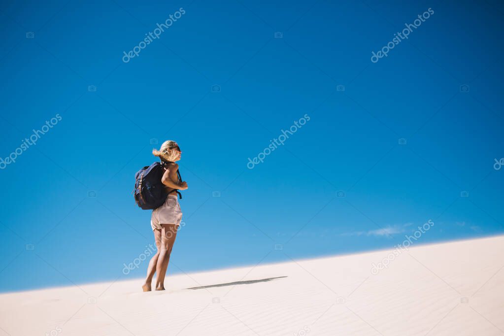 Back view of barefoot blonde woman with backpack standing on sandy dune and looking away against clear blue sky in sunlight 