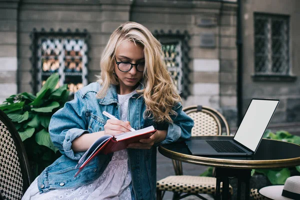 Concentrated young student in spectacles making notes in notebook while leaning on cafe table with laptop with empty screen on city street