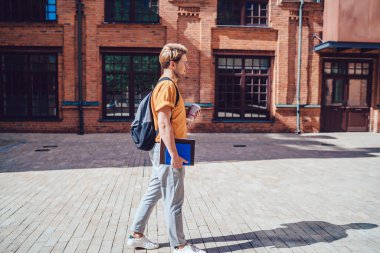 Gündüz giyimli, evrak işleriyle dolu gözlüklü, sırt çantalı, güneşli bir günde sokakta kahve içen yakışıklı genç adamın yan görüntüsü.