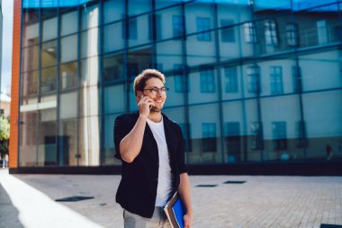 Gözlüklü, beyaz tişörtlü, koyu renk ceketli neşeli bir adam. Şehirde elinde not defterleriyle dikilip gözlerini kaçırırken cep telefonuyla sohbet ediyor.