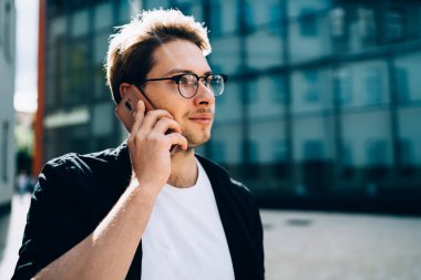 Gözlüklü, kendine güvenen, aktif bir erkek. Şehir merkezinde durup başka yöne bakarken cep telefonuyla bilgi dinliyor.