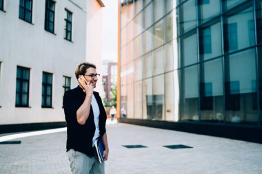 Modern şehir caddesinde güneşli bir günde kaldırımda dikilirken gülümseyen ve telefonlara cevap veren mutlu genç adam.