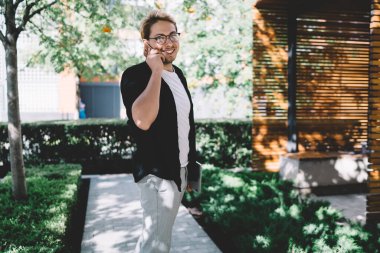 Moda gözlüklü, siyah gömlekli, akıllı telefondan konuşan ve şehir meydanında dizüstü bilgisayarla beklerken harika haberler alan başarılı genç girişimcinin yan görüntüsü. 