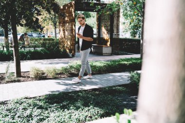 Yaz günü şehir parkında asfalt yolda yürürken sosyal ağda cep telefonu kullanan ve haberleri okuyan genç bir adamın yan görüntüsü 