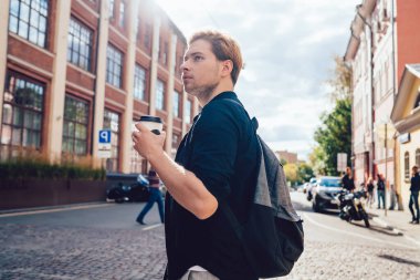 Sırt çantalı, rahat giyimli, şehirde bir fincan kahve içen arka tarafa aydınlatılmış dikkatli genç bir adamdan.