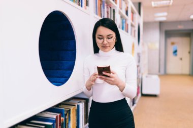 Gözlüklü güzel Asyalı kız öğrenci modern kütüphanede kitaplığın yanında dururken cep telefonuyla cep telefonu ekranına bakıyor.