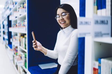 Kütüphanede kitaplıkların arasında oturan ve cep telefonu kullanan, gözlüklü, mutlu, gülümseyen genç bir kadının yan görüntüsü