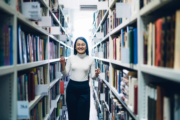 身穿白衬衫和黑裙的年轻亚洲女学生兴高采烈地站在高校图书馆的白书架之间看书 — 图库照片