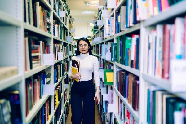 亚洲黑发女子 戴着白色高领眼镜 蓝色长裤 手里拿着五颜六色的书 走在图书馆的书架上 — 图库照片
