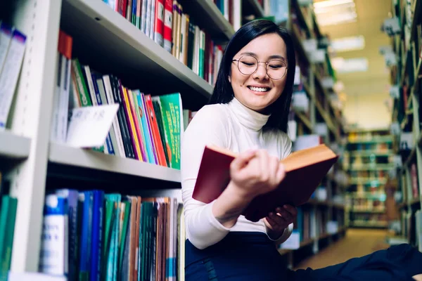 年轻漂亮的亚洲女性 戴着圆圆的眼镜 面带微笑地坐在书架旁边的图书馆地板上看书 — 图库照片