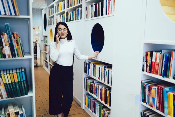 Thoughtful stylish Asian woman in white turtleneck talking on mobile phone and leaning on round recess in designed bookshelves in light library