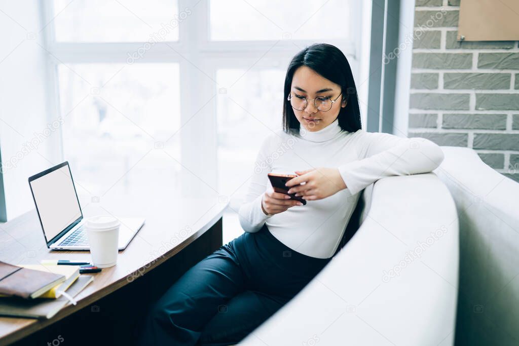 Long haired clever Asian woman in glasses and white turtleneck surfing mobile phone leaning  on back of sofa at table with open laptop 