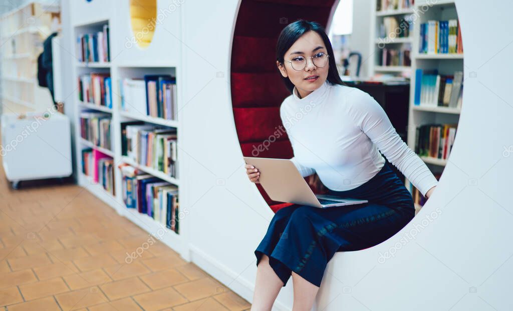 Young curious Asian woman with laptop on knees sitting on round bench in contemporary library unit with bookshelves while leaning forward and looking away