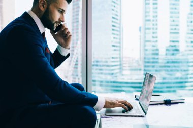 Koyu renk takım elbiseli ekin yöneticisinin yan görünümü bilgisayarını karıştırıyor ve çağdaş ofiste masanın yanında otururken akıllı telefondan konuşuyor.