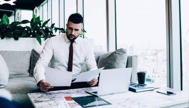 İş projesinde çalışırken belgelere bakan düşünceli bir erkek yemlik. Kafede dizüstü bilgisayarla rahat bir koltukta oturuyor.