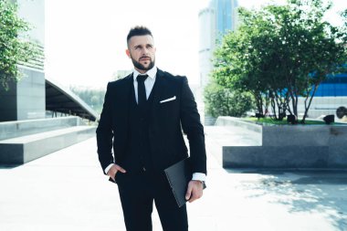 Siyah takım elbiseli düşünceli genç bir adam. Yanında not defteri ve el ele. Güneşli bir günde modern şehrin caddesinde buluşmayı bekliyor.