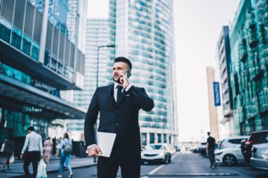 Şehir caddesindeki modern binaların ortasında akıllı telefondan konuşurken elinde tablet taşıyan ve bakışlarını çeviren koyu renk takım elbiseli zeki adam.