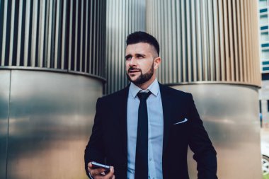 Resmi sakallı iş adamı akıllı telefon taşıyor ve modern şehrin merkezindeki çağdaş binanın önünde dikilirken başka yere bakıyor.