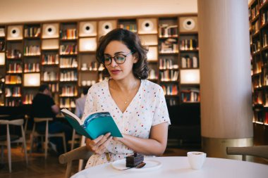 Açık renk yazlık elbiseli ve gözlüklü güzel bir kadın ilginç bir kitap okurken çikolatalı kek ve kahveyle modaya uygun bir kafede oturuyor.