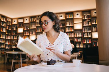 Masada ve şık kafeteryada bir fincan kahve ve bir parça kekle otururken gündelik giysiler ve gözlük okuyan zeki bayan öğrenci.