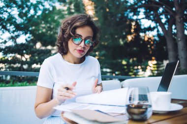 Beyaz tişörtlü ve gözlüklü bir kız öğrenci gün ışığında kafede masada otururken üniversite ödevlerini yapıyor.