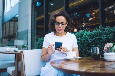 Gözlüklü düşünceli genç bir kadın teras kafedeki masada rahat bir sandalyede dinlenirken cep telefonuyla mesajlaşıyor ve içki içiyor.