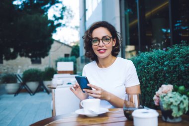 Kıvırcık saçlı, gülümseyen genç bir kadın kameraya bakıyor ve akıllı telefonlara bakarken yeşil bitkilerin yanındaki masada oturuyor.