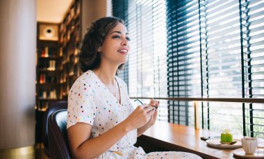 Zarif yaz elbisesi içinde neşeli genç bir kadın rahat bir kafede oturuyor ve elinde bardaklarla pencereye bakıyor.