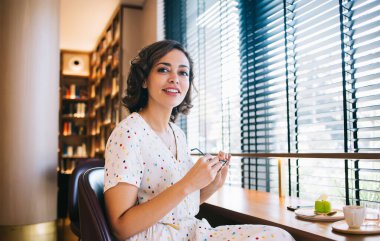 Gözlüklü ve beyaz elbiseli gülümseyen genç bir kadın sıcak kafeteryada bir fincan kahve ve tatlıyla masada oturuyor ve kameraya bakıyor.