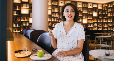 Günümüz kafeteryasında kitaplıklarla oturmuş kahve içen gündelik kıyafetli düşünceli genç bir kadın.