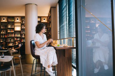 Gündelik kıyafetlerle gezen genç bir kadın pencerenin yanında kahve ve tatlıyla birlikte kitap rafları ile modern kafede oturuyor.