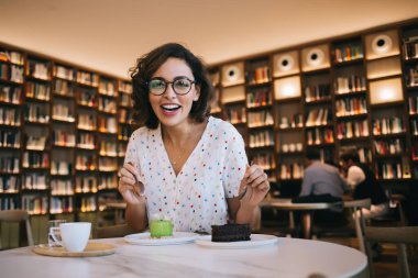 Gözlüklü ve günlük kıyafetleriyle gülümseyen genç bir kadın zevkli tatlılar deneyip moda kafede otururken kameraya bakıyor.