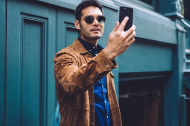 Sırt çantalı, şık giyinmiş ve güneş gözlüklü yakışıklı genç bir erkek. New York caddesinin mavi binasının karşısında dikilirken selfie çekiyor.