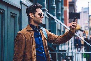 Sırt çantalı ve gözlüklü yakışıklı genç adam New York caddesinin mavi binasında dururken gülümsüyor ve selfie çekiyor.