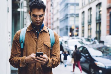Gözlüklü, kahverengi süet ceketli, sırt çantalı ve kulaklıklı, New York 'ta ofis binalarının arkasındaki caddede duran akıllı telefonlu yakışıklı bir erkek.